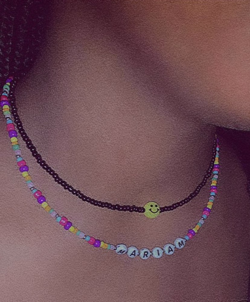 Close-up of neck with two beaded necklaces, one colorful and one spelling out ‘MARIAM’ with a smiley face bead in the center